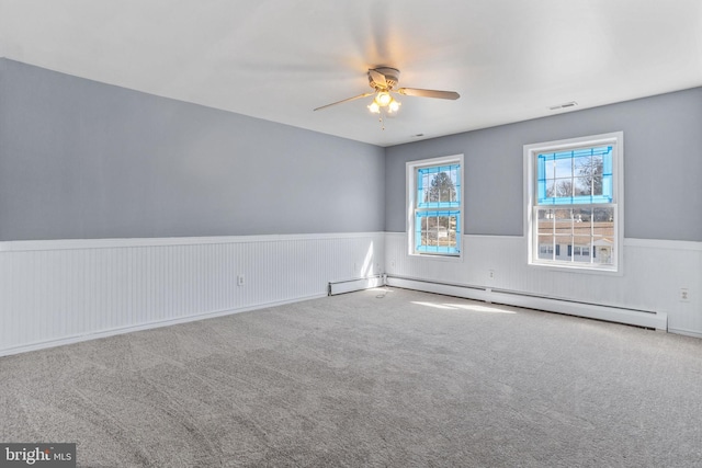 carpeted empty room featuring ceiling fan, visible vents, baseboard heating, and wainscoting