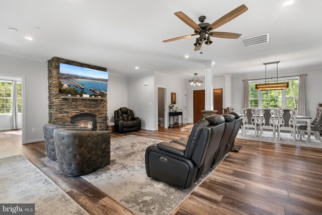 living room featuring wood-type flooring, ceiling fan with notable chandelier, a stone fireplace, ornamental molding, and ornate columns