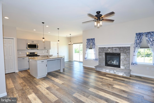 kitchen featuring a fireplace, appliances with stainless steel finishes, an island with sink, ceiling fan, and light stone counters