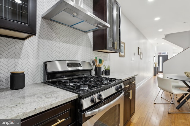 kitchen featuring light stone counters, dark brown cabinets, wall chimney range hood, light hardwood / wood-style floors, and gas stove