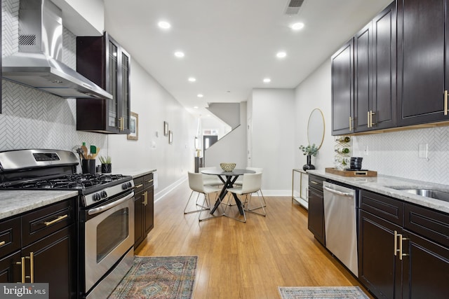 kitchen featuring light wood-type flooring, tasteful backsplash, wall chimney range hood, appliances with stainless steel finishes, and light stone countertops