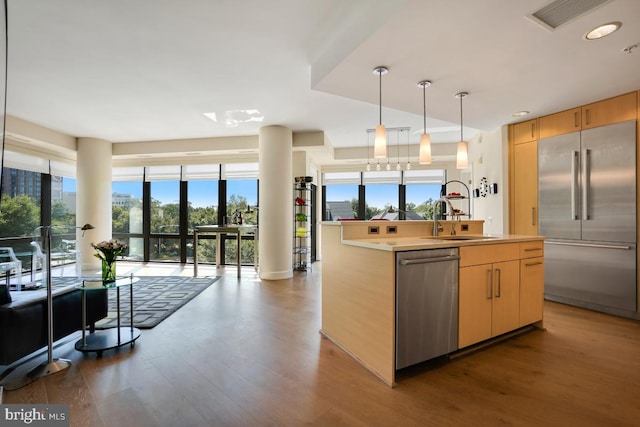 kitchen with light brown cabinets, a center island with sink, hanging light fixtures, sink, and appliances with stainless steel finishes