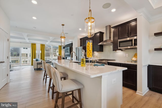 kitchen featuring a center island with sink, wall chimney exhaust hood, light wood-type flooring, decorative light fixtures, and stainless steel appliances