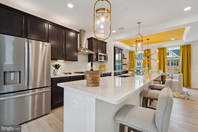 kitchen featuring wall chimney exhaust hood, stainless steel appliances, light hardwood / wood-style flooring, hanging light fixtures, and a breakfast bar area