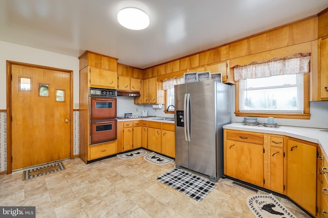 kitchen featuring a sink, light countertops, stainless steel refrigerator with ice dispenser, under cabinet range hood, and double oven