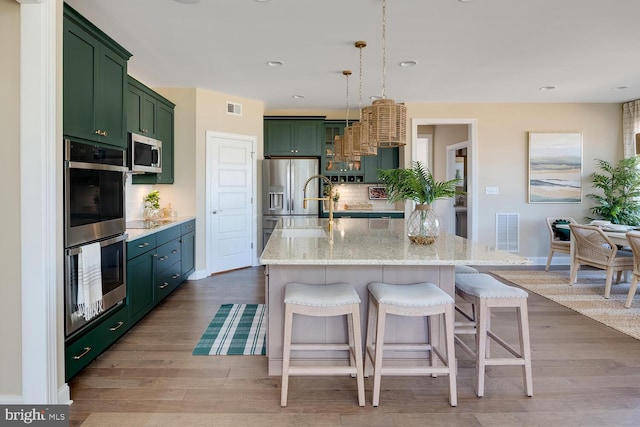 kitchen featuring hanging light fixtures, appliances with stainless steel finishes, light stone counters, light hardwood / wood-style floors, and an island with sink