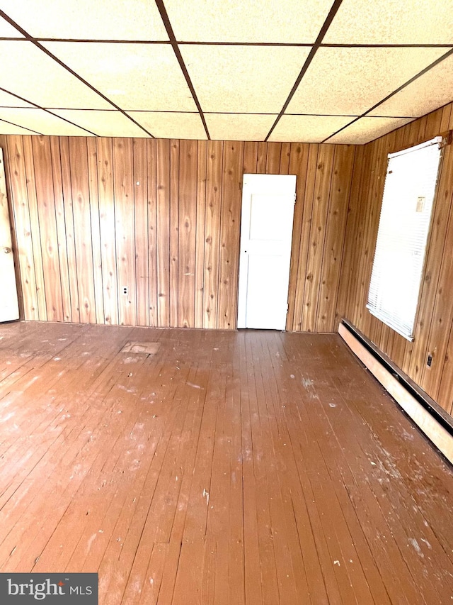 empty room with a baseboard heating unit, wood walls, wood-type flooring, and a drop ceiling