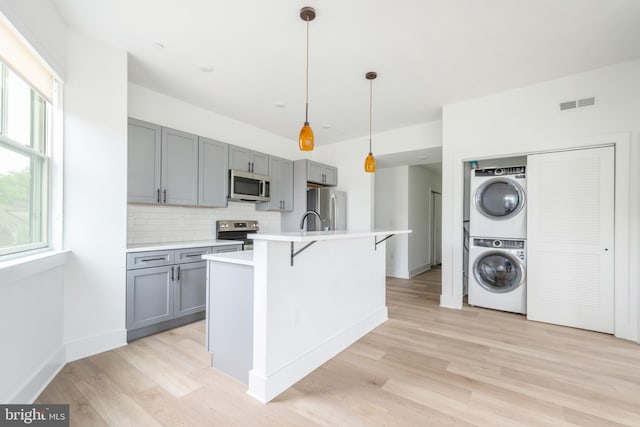 kitchen with pendant lighting, light hardwood / wood-style flooring, stacked washer / drying machine, appliances with stainless steel finishes, and a center island with sink