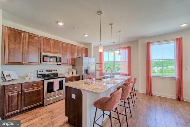 kitchen featuring pendant lighting, appliances with stainless steel finishes, an island with sink, light stone counters, and light wood-type flooring