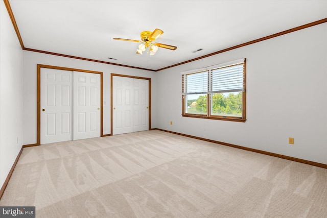 unfurnished bedroom with ornamental molding, two closets, ceiling fan, and light colored carpet