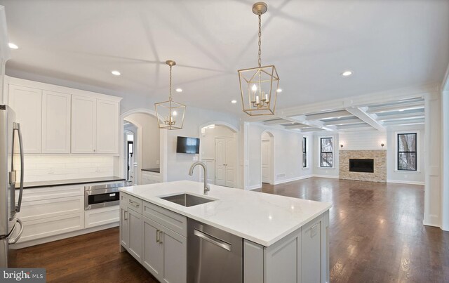 kitchen with hanging light fixtures, a stone fireplace, stainless steel appliances, a kitchen island with sink, and sink