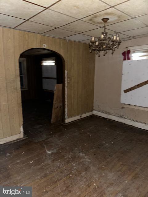 spare room with a notable chandelier, wooden walls, a drop ceiling, and dark wood-type flooring