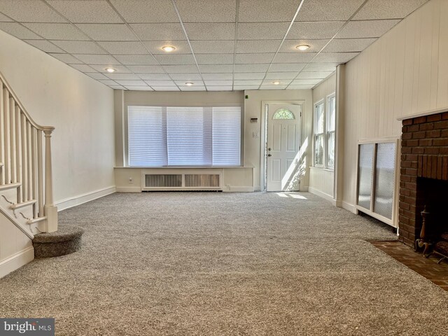 unfurnished living room featuring a drop ceiling, dark colored carpet, and a brick fireplace