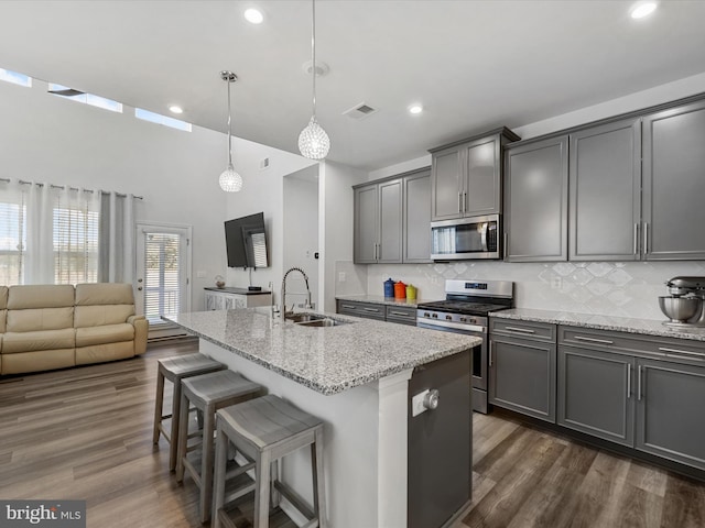 kitchen with light stone counters, stainless steel appliances, sink, hanging light fixtures, and an island with sink