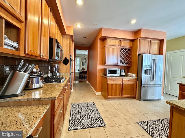 kitchen with light tile patterned floors, stainless steel appliances, backsplash, brown cabinets, and light stone countertops