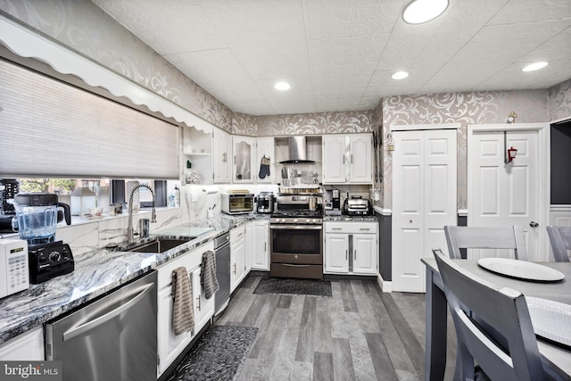 kitchen with a sink, white cabinetry, appliances with stainless steel finishes, wall chimney exhaust hood, and wallpapered walls