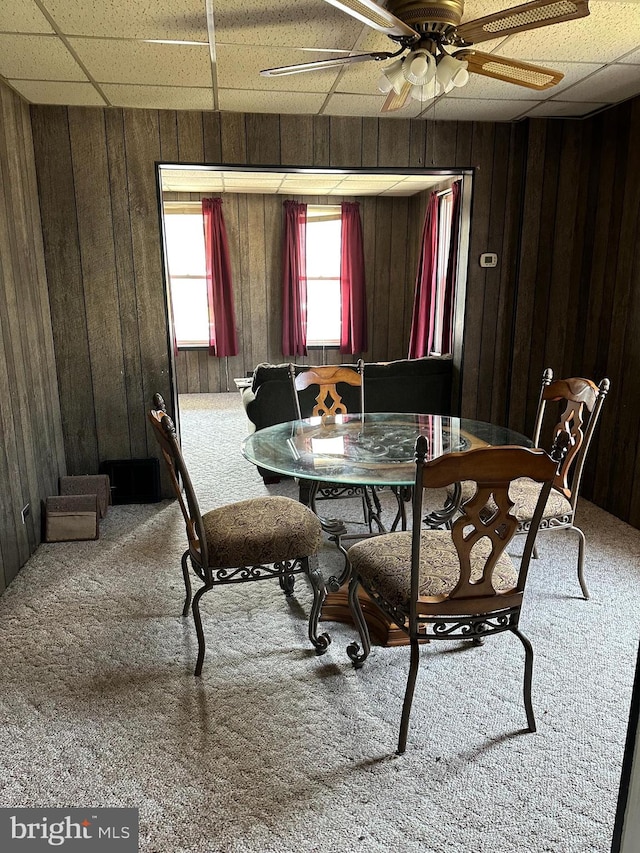 dining room featuring carpet, ceiling fan, wood walls, and a drop ceiling