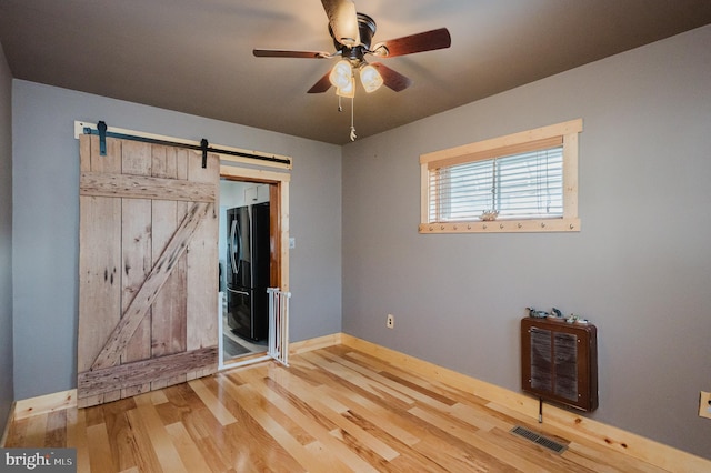 unfurnished bedroom featuring black fridge, hardwood / wood-style flooring, ceiling fan, a barn door, and heating unit