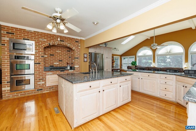 kitchen with light wood-type flooring, appliances with stainless steel finishes, vaulted ceiling with skylight, ceiling fan, and a kitchen island with sink