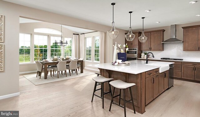 kitchen featuring light wood-type flooring, wall chimney exhaust hood, a kitchen island with sink, and a notable chandelier