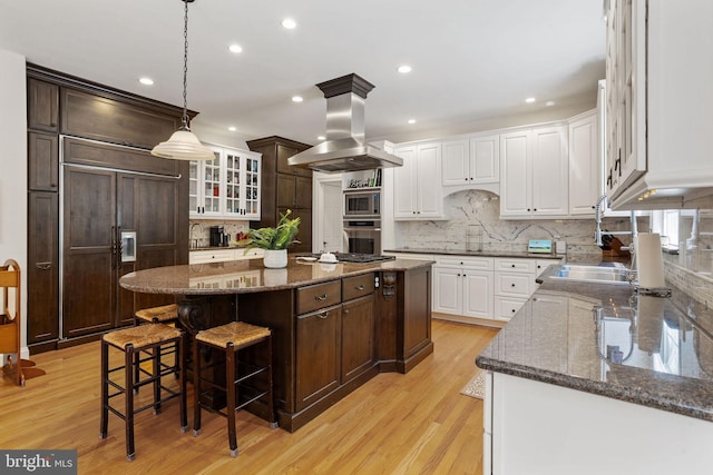 kitchen with dark stone countertops, light hardwood / wood-style flooring, built in appliances, a kitchen island, and island range hood