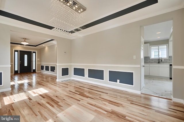 unfurnished living room with ceiling fan with notable chandelier, a raised ceiling, crown molding, sink, and light hardwood / wood-style flooring