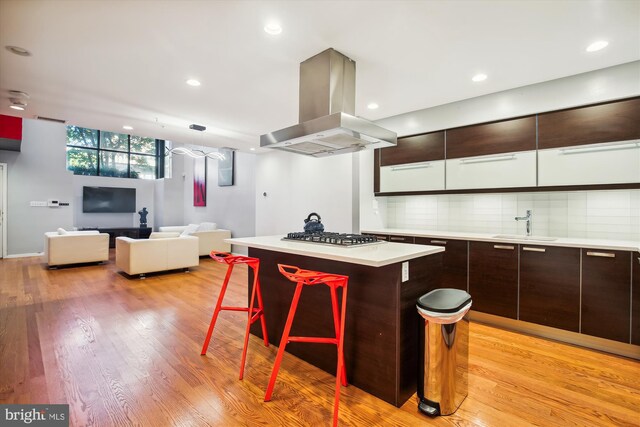 kitchen with a kitchen island, light hardwood / wood-style flooring, stainless steel gas cooktop, island exhaust hood, and a breakfast bar