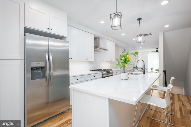 kitchen with appliances with stainless steel finishes, light hardwood / wood-style floors, white cabinetry, wall chimney exhaust hood, and a kitchen island with sink