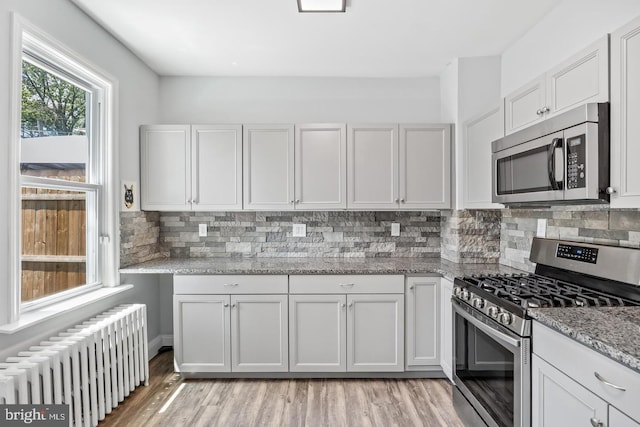 kitchen featuring light hardwood / wood-style flooring, stainless steel appliances, radiator heating unit, light stone countertops, and decorative backsplash