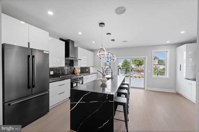 kitchen with stainless steel appliances, an island with sink, hanging light fixtures, white cabinets, and wall chimney range hood