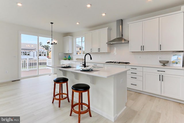 kitchen with an island with sink, sink, wall chimney exhaust hood, white cabinetry, and light hardwood / wood-style floors