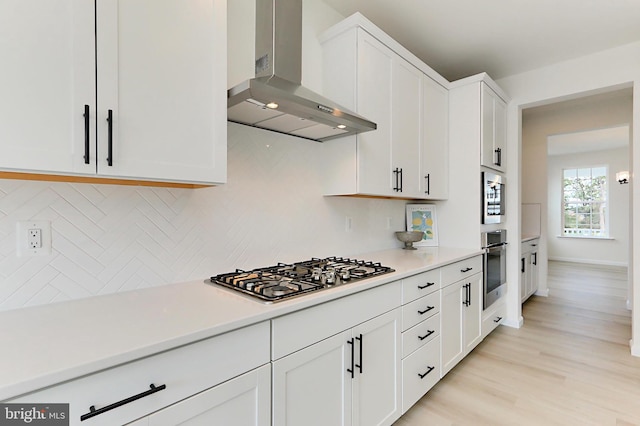 kitchen with tasteful backsplash, white cabinets, stainless steel appliances, light wood-type flooring, and wall chimney range hood