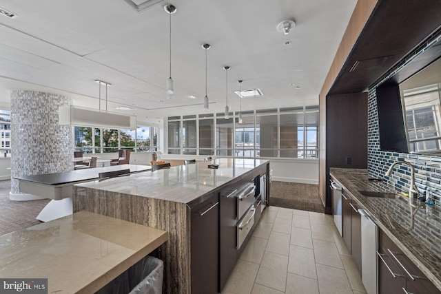 kitchen featuring hanging light fixtures, light stone countertops, a spacious island, dark brown cabinets, and decorative backsplash