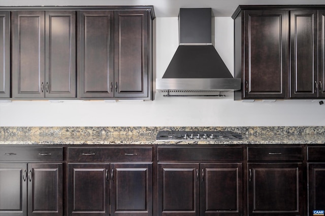 kitchen featuring stainless steel gas stovetop, light stone counters, dark brown cabinetry, and wall chimney range hood