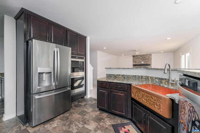 kitchen with dark brown cabinets, stainless steel appliances, light stone countertops, ceiling fan, and a stone fireplace