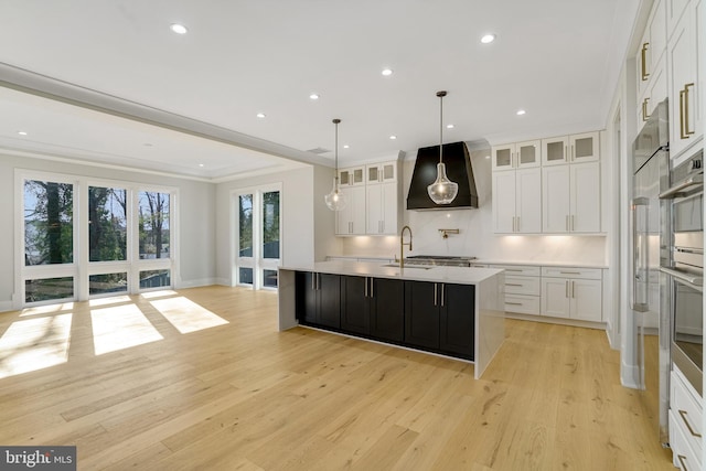 kitchen with pendant lighting, white cabinets, light wood-type flooring, an island with sink, and custom range hood