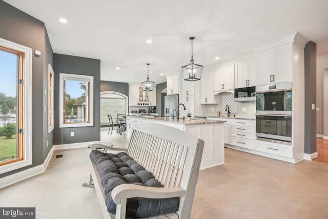 kitchen with white cabinets, an island with sink, double oven, and hanging light fixtures