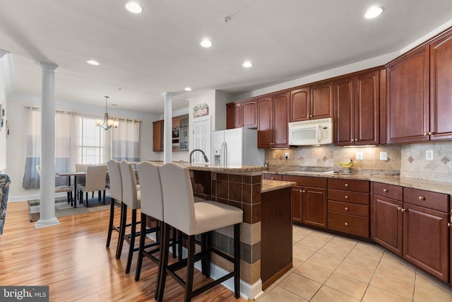 kitchen featuring white appliances, decorative light fixtures, a center island with sink, decorative columns, and a breakfast bar area