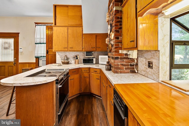 kitchen with wallpapered walls, brown cabinets, a peninsula, dark wood-style floors, and black appliances