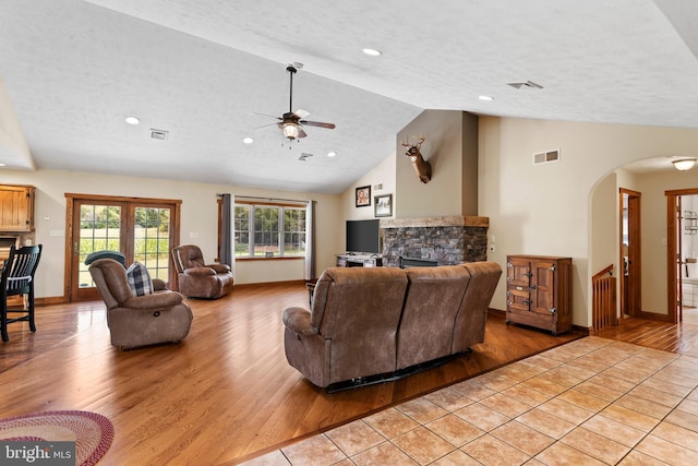 living room featuring light wood-type flooring, a textured ceiling, high vaulted ceiling, a stone fireplace, and ceiling fan