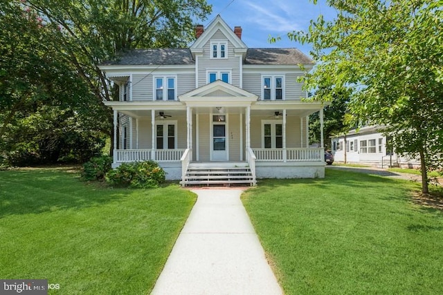 victorian house featuring a porch, a ceiling fan, a front lawn, and a chimney