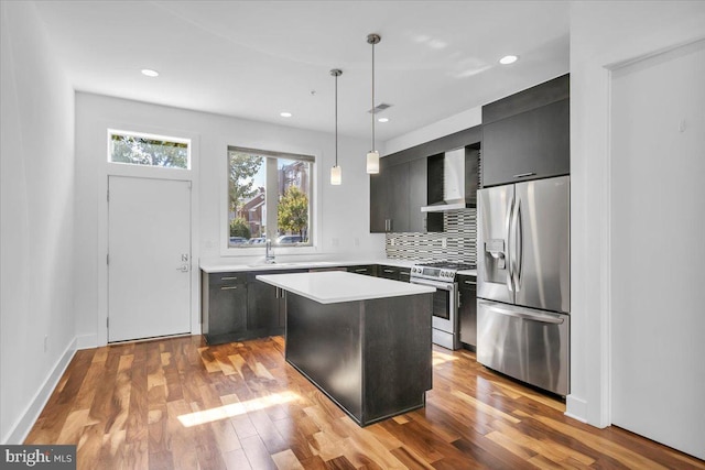 kitchen with wall chimney range hood, light wood-type flooring, a kitchen island, stainless steel appliances, and hanging light fixtures