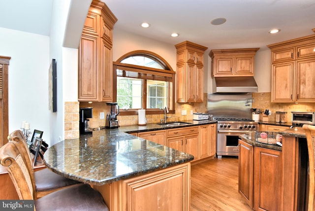 kitchen featuring sink, high end stainless steel range, exhaust hood, dark stone countertops, and light wood-type flooring