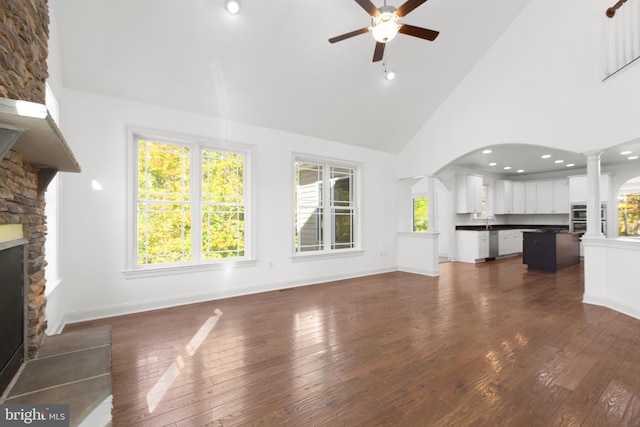 unfurnished living room with a stone fireplace, dark hardwood / wood-style flooring, decorative columns, high vaulted ceiling, and ceiling fan