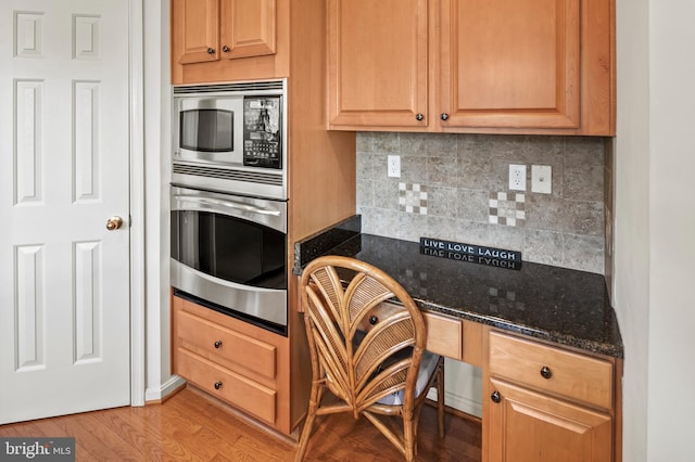 kitchen featuring built in desk, light hardwood / wood-style flooring, dark stone countertops, appliances with stainless steel finishes, and tasteful backsplash