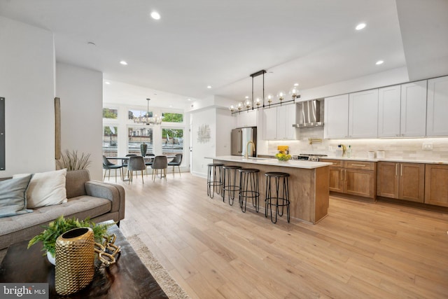 kitchen featuring a large island with sink, an inviting chandelier, light hardwood / wood-style flooring, decorative light fixtures, and wall chimney range hood