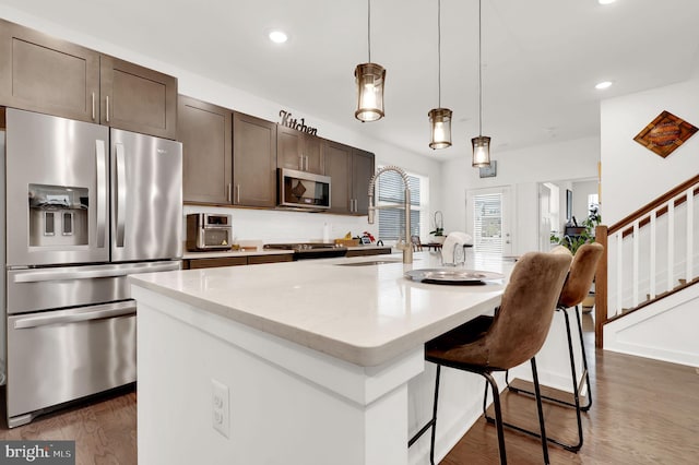 kitchen with appliances with stainless steel finishes, dark hardwood / wood-style flooring, pendant lighting, dark brown cabinetry, and a center island with sink