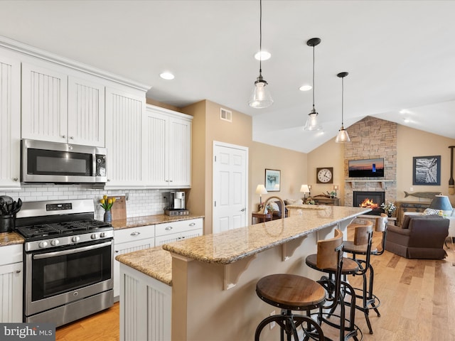 kitchen featuring white cabinets, an island with sink, appliances with stainless steel finishes, a fireplace, and vaulted ceiling