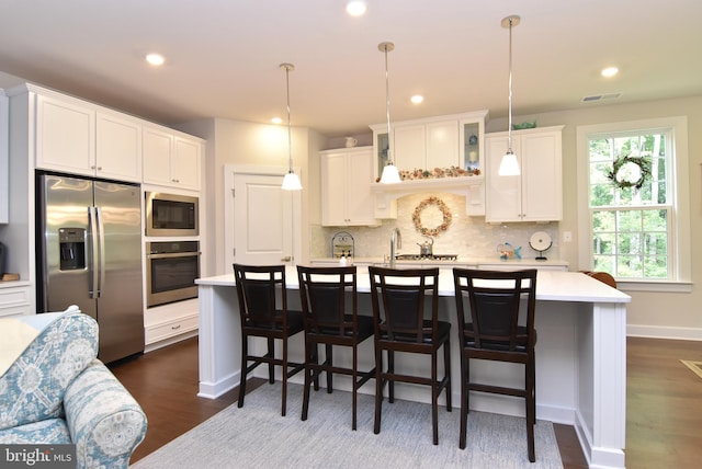 kitchen featuring dark wood-type flooring, a kitchen island with sink, white cabinetry, appliances with stainless steel finishes, and decorative light fixtures