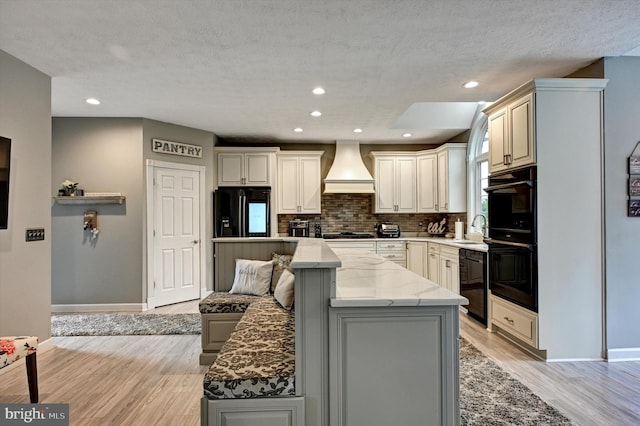 kitchen featuring black appliances, light wood-type flooring, a center island, custom exhaust hood, and light stone counters